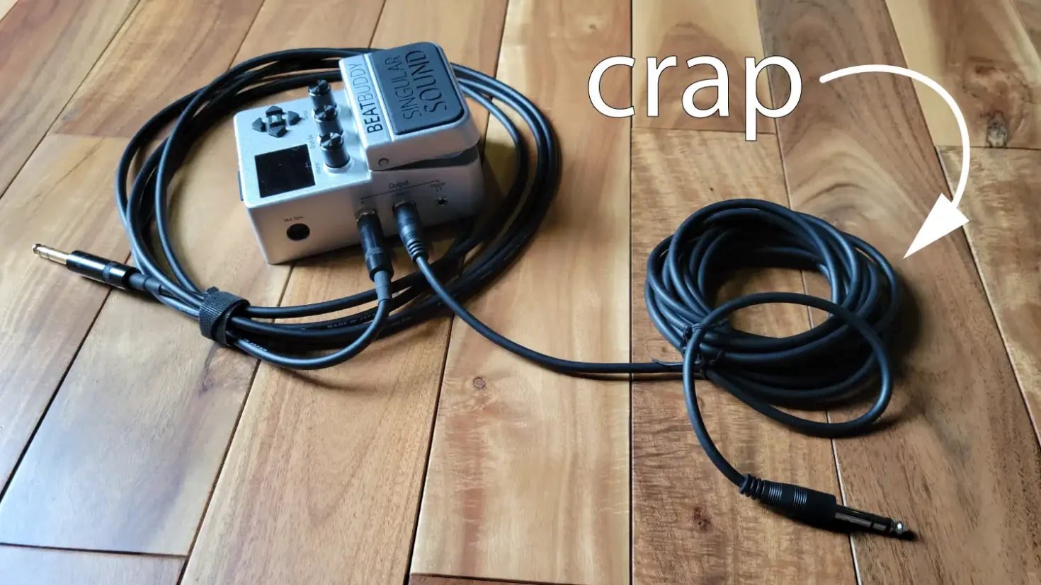 BeatBuddy and two cables plugged into it, over a wooden floor with text "crap" and an arrow pointing to one of the cables.